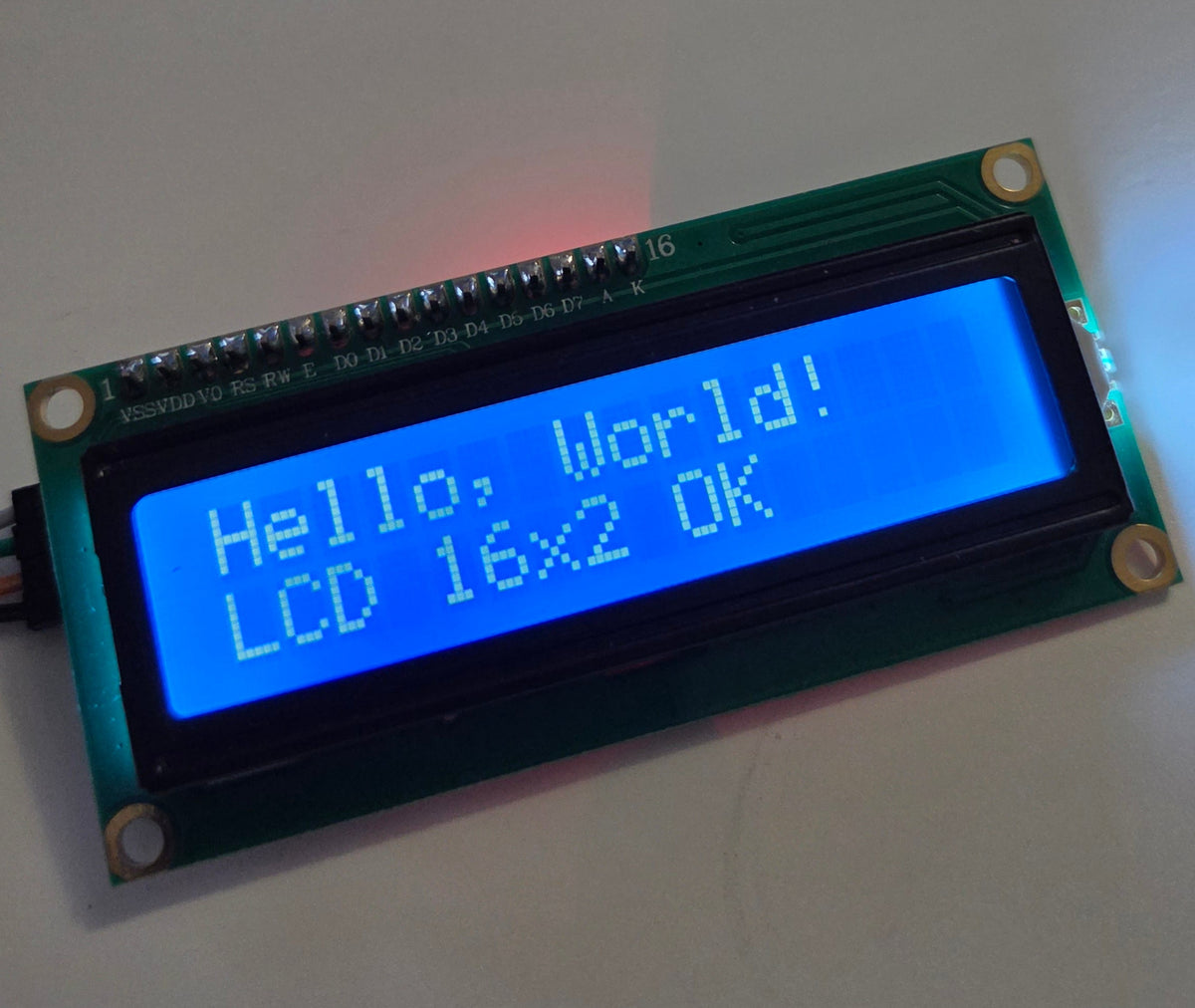 A small LCD 16x2 display module on a green circuit board, showing a blue screen with the text 'Hello, World! LCD 16x2 OK' on a light gray surface.