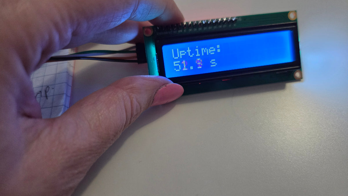 A close-up of a hand holding a small LCD display module with a blue screen showing uptime text, set against a plain light background.