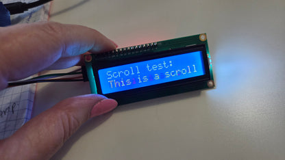 A hand holding a small LCD display module with a green circuit board and a blue backlit screen showing the text 'Scroll test: This is a scroll'.