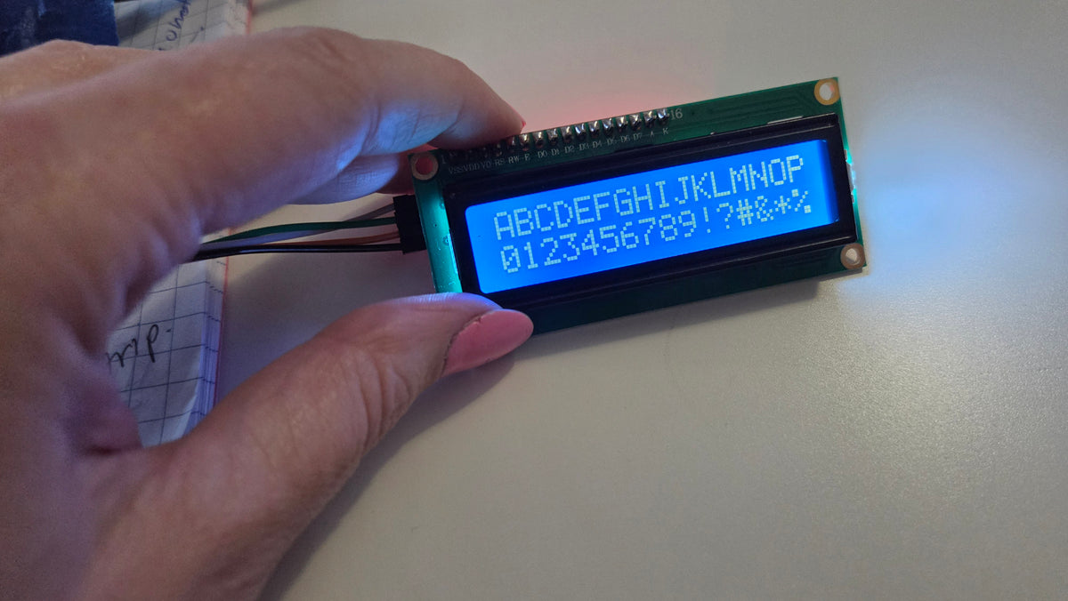 A close-up of a hand holding a small green LCD display module with a blue backlit screen showing white characters, against a light tabletop background.