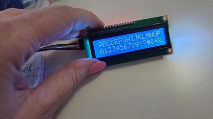 A close-up of a hand holding a small green LCD display module with a blue backlit screen showing white characters, against a light tabletop background.