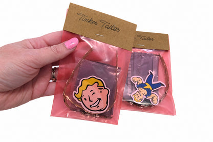 A hand with pink nail polish holds two small packaged items on a white surface. The items appear to be novelty glasses or cup-shaped accessories with Fallout-style character graphics, each wrapped in a translucent pink pouch with a brown paper tag.